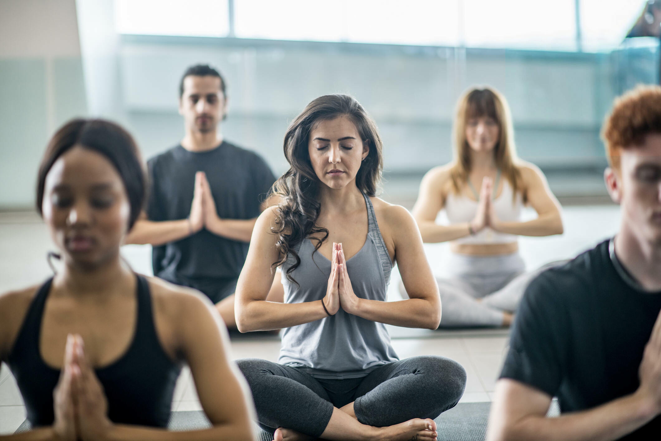 A multi-ethnic group of adults are indoors in a fitness center. They are sitting on the floor and meditating with their legs crossed. One woman is smiling at the camera.