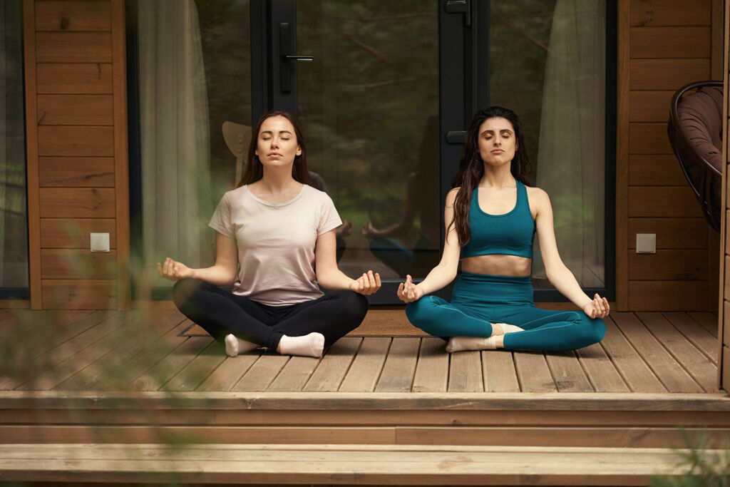 Young female friends sitting on floor in the lotus pose and meditate, relaxing outdoors