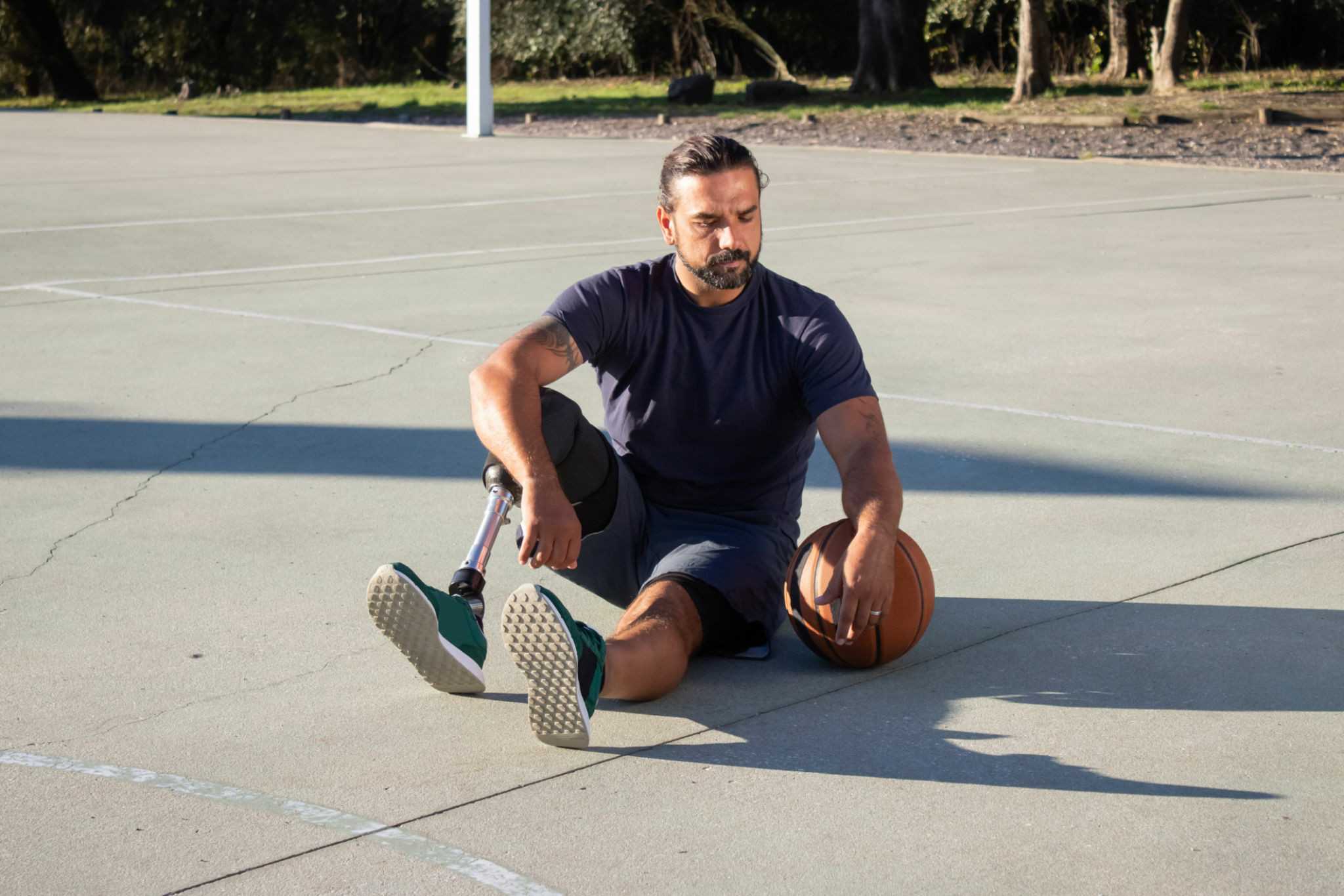 Bearded middle-aged man with prosthesis sitting at sports ground while holding a ball beside and looking at it. Man with disability having break during ball game. Disability, active lifestyle concept